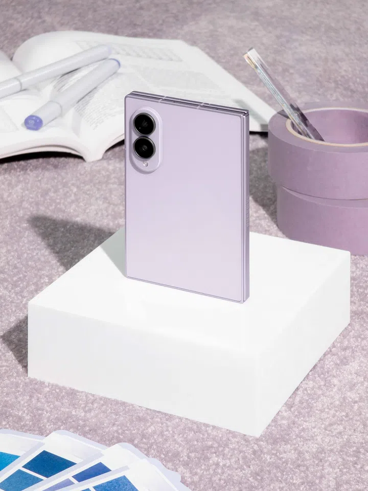 Samsung Galaxy Z Fold8 Lavender（薰衣草紫）产品展示图。手机以折叠状态直立摆放于白色展示台上，展示雾面淡紫色背板及双镜头相机模组，整体机身线条简洁俐落。背景摆放打开的书本、紫色马克笔、紫色胶带及设计工具，营造创意工作室氛围。柔和的自然光线搭配浅紫色背景，突显Galaxy Z Fold8轻薄设计、精致质感及适合内容创作与日常办公的使用情境。