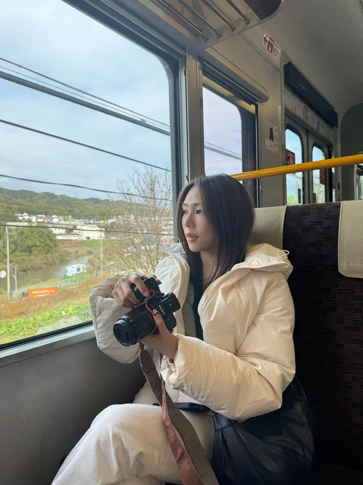 Rachel Wan坐在行驶中的列车靠窗座位上，身穿米白色羽绒外套，手持相机望向窗外风景，窗外可见郊外山景与河流，画面展现旅途中的安静与专注时刻。Rachel Wan：最好的感情，是让我更像自己