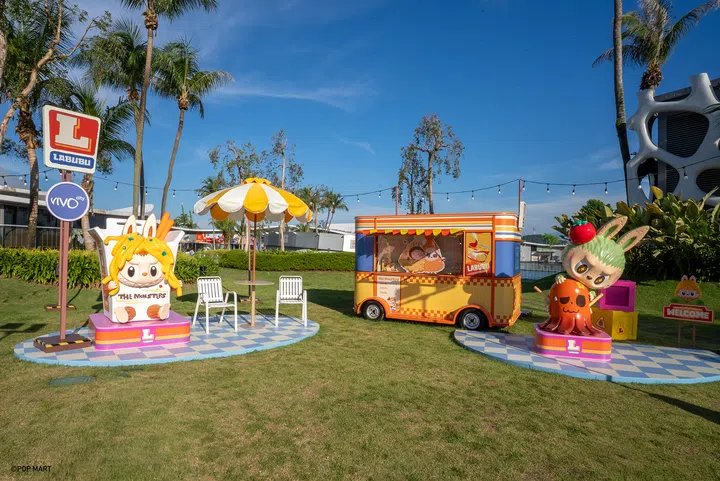 照片拍摄于VivoCity天台，画面中央有LABUBU主题打卡区，包含黄色与橙色相间的小餐车、巨型LABUBU角色雕塑、遮阳伞与座椅，背景是蓝天与棕榈树，整体氛围明亮俏皮，充满欢乐气息。
