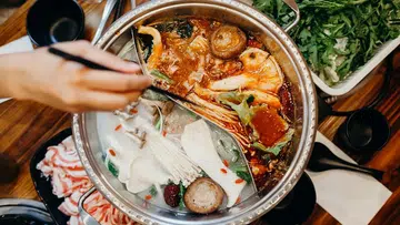 一张从上方俯拍的火锅用餐画面：圆形鸳鸯火锅被金属隔板分成两侧，一边是红色麻辣汤底，汤面可见辣椒、香料、虾、贝类和蔬菜；另一边是清汤或牛奶汤底，放有金针菇、香菇、枸杞、青菜等配料。画面左侧有一只手正使用汤勺在锅中翻动食材，增添用餐动态感。火锅周围摆放着多盘配料，包括切片肉类、绿叶蔬菜与调味碗，桌面为木质纹理，整体呈现热气腾腾、丰盛且具有聚餐氛围的亚洲火锅场景。