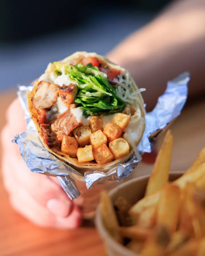 手持一份现做的凯撒风味Cali Burrito卷饼，饼内包裹炭火烤鸡肉块、金黄酥脆的薯块、新鲜罗马生菜、番茄丁与凯撒酱，食材层层堆叠，口感丰富饱满。卷饼外层以银色锡纸包裹，旁边搭配一份金黄色薯条，呈现适合新春期间轻松享用的异国风味美食选择。新加坡大年初一初二照常营业餐厅推荐：8家不打烊好去处（2026马年）