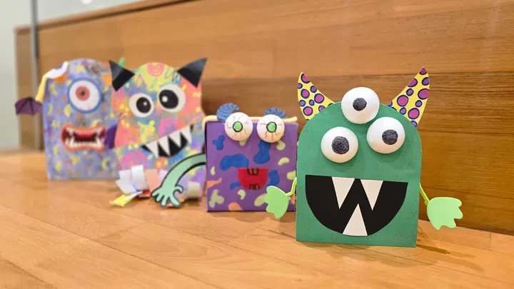 一排由儿童亲手制作、色彩缤纷的纸艺怪兽摆放在木地板上，展现创意与节日乐趣。新加坡万圣节去哪里玩推荐。