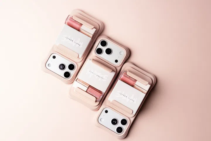 Rihanna创立的Fenty Beauty与科技配饰品牌CASETiFY推出限定联名系列，图片以柔和的裸粉色为背景，三部配备不同尺寸镜头模组的智能手机斜向排列，展示Cocoa Nude裸棕粉色手机壳与On The Go Essentials MagSafe 2-in-1 Holder。手机背面装有白色Fenty Beauty品牌蜜粉盒及粉棕色Gloss Bomb唇蜜，利用MagSafe磁吸配件固定在手机壳上，呈现将化妆品与手机结合的创新设计。整体以裸粉、奶油白及柔和玫瑰色为主调，突出Fenty Beauty与CASETiFY兼具美感及实用性的随身补妆概念。