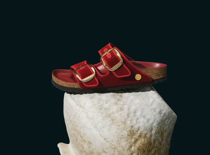 图片展示一双红色Birkenstock Arizona凉鞋，放置在浅色天然石块造型的展示台上。鞋款采用经典双带设计，鞋面为柔软的红色天鹅绒材质，搭配醒目的金色金属大扣环，整体外观时尚而富有节庆气息。鞋床呈现深红色调，侧边可见天然软木鞋底结构与黑色防滑橡胶底，展现品牌标志性的舒适与稳固设计。画面背景为深色调，凸显鞋款的鲜艳红色与精致细节，营造简约又具质感的视觉风格，非常适合作为新年穿搭的舒适鞋款选择；2026新年鞋推荐：舒适好穿又好看的拜年鞋款。