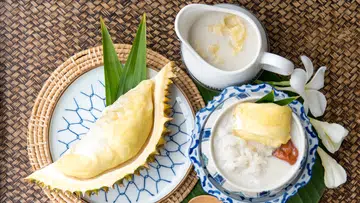 一盘榴莲与泰式甜品摆放在藤编桌布上，包括榴莲果肉、椰浆糯米与装有榴莲酱的白色小壶，旁边点缀着热带绿叶和白色鸡蛋花。