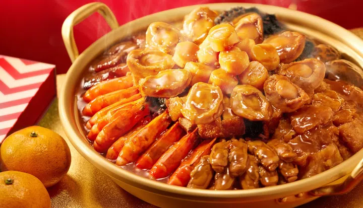 金色砂锅中盛装粤式风味盆菜，整齐铺排整虾、鲍鱼、花胶、干贝与多款海味与配料，表面淋上浓郁酱汁并冒着热气，旁边点缀柑橘与新年礼盒装饰，营造喜庆温暖的农历新年团圆饭氛围，新加坡盆菜推荐2026 农历新年必吃餐厅与价格整理。