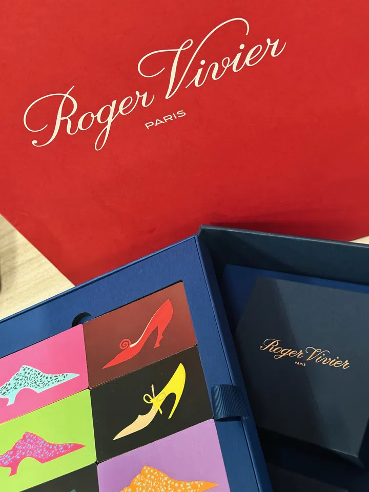 Roger Vivier中秋礼盒局部特写，背景为印有浅金色品牌手写体标志的红色外包装，前景打开的深蓝色礼盒内排列多张彩色鞋履图案卡片。桃粉、鲜绿、淡紫及深色底面上，分别绘有尖头高跟鞋与镂空短靴，呈现蝴蝶结、弧形鞋跟等细节。右侧放置带金色品牌字样的深蓝色封套，中央可见蓝色织带拉手。