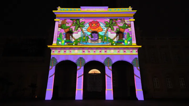 National Museum of Singapore外墙夜间上演大型投影光雕秀，建筑立面披上鲜艳的紫色、粉色、黄色及绿色动画图案，中央可见以新加坡神话与民间传说为灵感设计的卡通狮子、热带植物、喷泉及飞鸟等元素，营造充满奇幻色彩的沉浸式视觉体验。整座历史建筑在灯光映照下焕然一新，展现Light Together Bras Basah.Bugis 2026期间融合艺术、文化与科技的夜间灯光装置。
