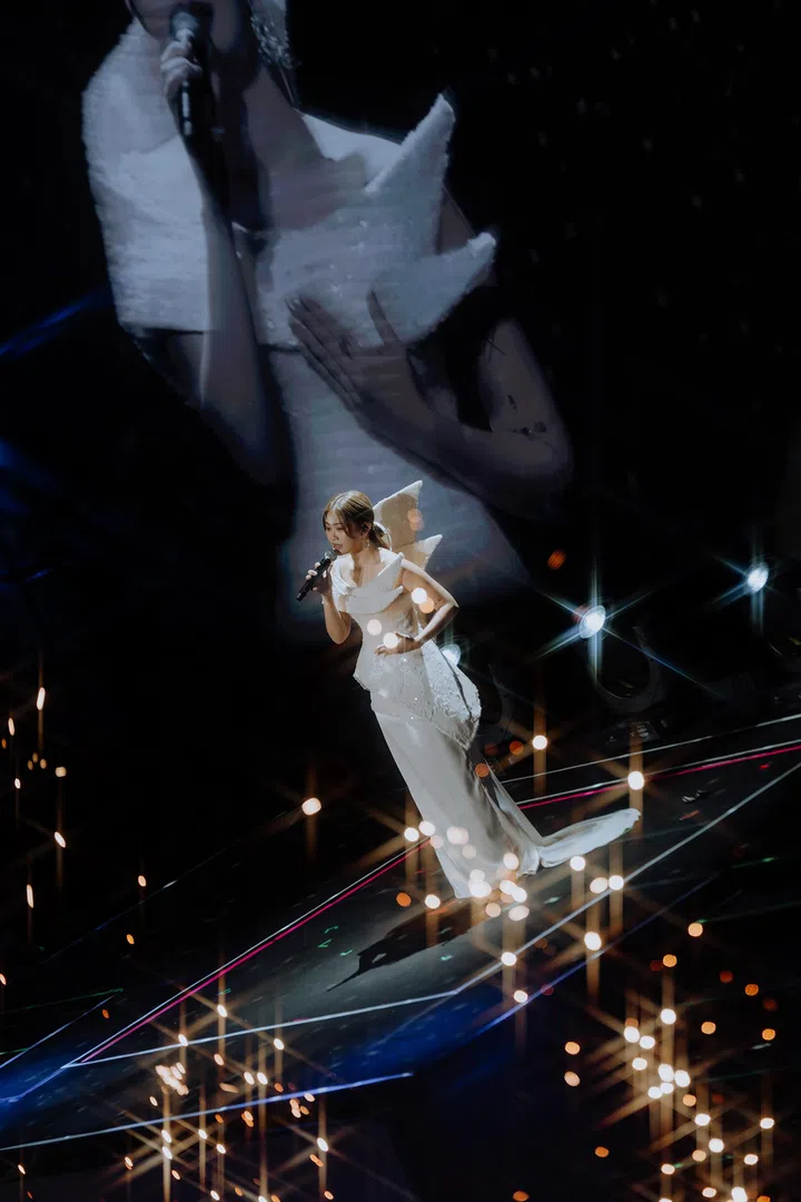 DIOR大颖在新加坡演唱会舞台上演唱，身穿白色立体剪裁礼服，站在延伸舞台中央，手持麦克风专注演出。舞台上点点灯光如星火般闪烁，营造梦幻氛围；背景巨型屏幕映出她的特写影像，与现场身影交叠，形成层次丰富的视觉效果，展现她细腻而真挚的情感表达。