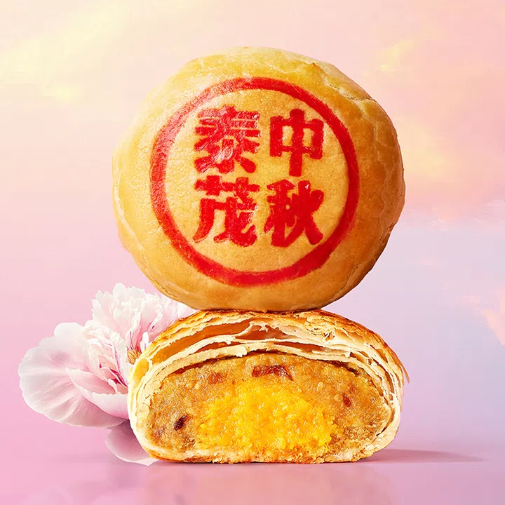 Thye Moh Chan泰茂栈咸豆沙蛋黄潮州月饼特写图，画面以粉色背景呈现一颗完整月饼与一颗切开的月饼。完整月饼表面印有红色“中秋献礼”字样；切开的月饼则清楚展现层层酥皮，以及咸豆沙与金黄色咸蛋黄内馅。旁边以浅粉色花朵点缀，整体突出潮州月饼酥松分层的外皮与丰富内馅。