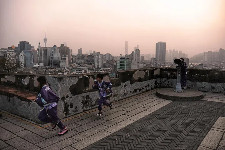 黄昏时分的城市观景平台上，三名身穿紫色运动服的孩子在围墙前奔跑玩耍，地面铺设为灰色石砖，形成规则的几何纹路。右侧设有固定式望远镜，面向远方的城市天际线。背景可见密集的高楼建筑与地标性高塔，在柔和的暮色与薄雾中呈现出层次分明的城市轮廓。孩子们的动作与城市静止的背景形成对比，画面捕捉日常生活中自然流动的瞬间与温暖气息。林道锦《谁在缝隙里种光》