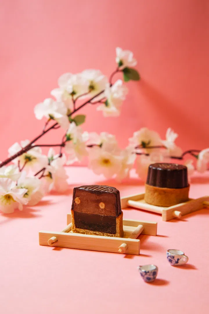 两枚 PAUL 巧克力月饼置于木质小托盘上，其中一枚切开，露出分层的巧克力馅料，深浅不一的可可色调清晰可见。背景点缀白色花枝与粉色底色，旁边摆放两只小巧的蓝白瓷茶杯，整体氛围甜美雅致。PAUL，2025中秋月饼必买推荐。