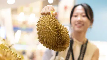 一位女子在榴莲摊前挑选榴莲，手中举着带刺的榴莲，背景是模糊的室内市场环境。