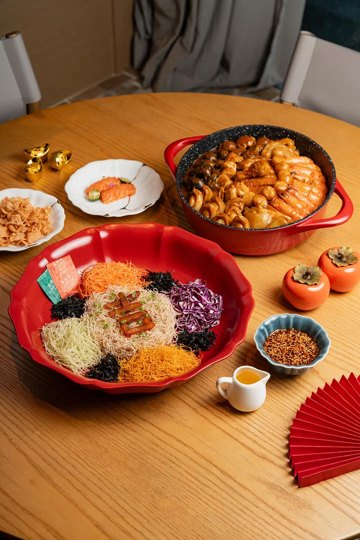 农历新年外带年菜与捞起鱼生组合展示图，木质圆桌上摆放一盘红色大碗盛装的捞起鱼生，配有白萝卜丝、红萝卜丝、紫甘蓝、海苔丝、脆饼及酱料包，中间放上叉烧片作为配料；后方是一锅丰盛盆菜，内含鲍鱼、海参、虾、香菇与卤肉等多种海味与肉类食材；桌上另有三文鱼片小碟、脆饼、酱料碗与酱汁壶，周围点缀金元宝装饰与红色扇形摆设，整体呈现适合家庭聚餐的新加坡农历新年外卖年菜与团圆饭氛围。