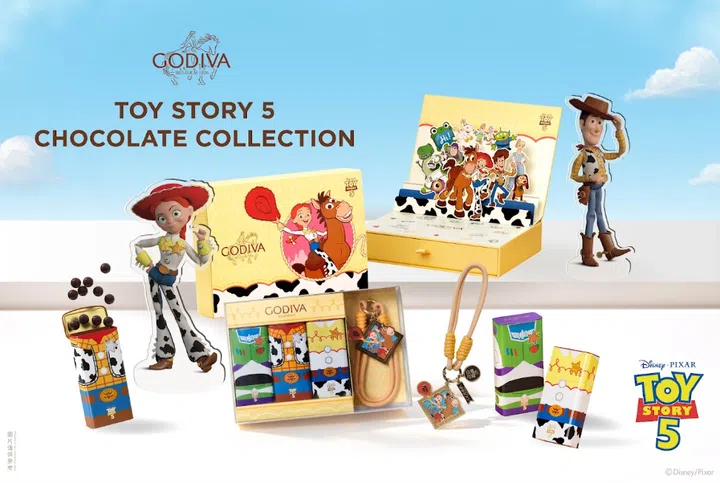 GODIVA x Disney and Pixar《Toy Story 5》联名巧克力系列宣传图，以蓝天白云为背景，中央陈列多款黄色主题礼盒、巧克力珍珠及收藏配件。画面左侧可见Jessie与Bullseye插画礼盒及Jessie人物立牌，右侧则有Woody人物立牌；后方打开的立体礼盒集合Woody、Jessie、Buzz Lightyear、三眼仔、Rex等多位《Toy Story》角色。前方展示三款分别以Buzz Lightyear、Woody及Jessie服装为灵感设计的巧克力珍珠包装，以及附有角色图案吊饰的钥匙扣与挂绳。画面上方印有GODIVA品牌标志及“TOY STORY 5 CHOCOLATE COLLECTION”字样，右下角则为Disney Pixar《Toy Story 5》标志，整体以黄色、蓝色及角色经典配色营造活泼的收藏氛围。