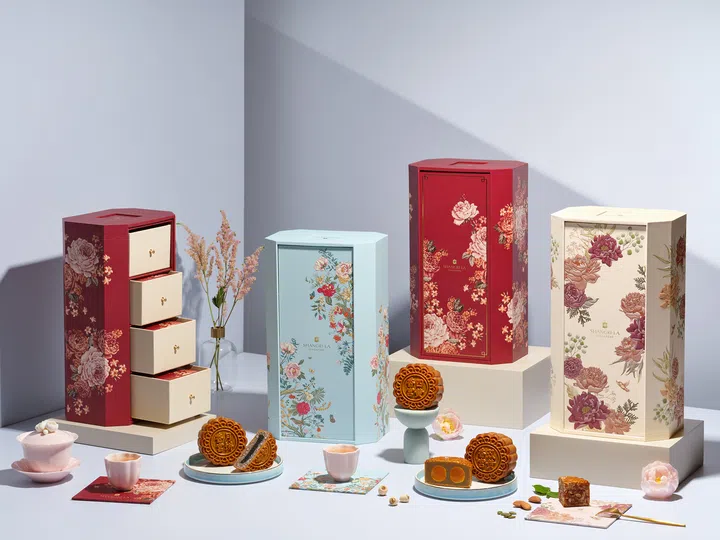 四款香格里拉酒店的中秋礼盒，分别以红、蓝、米白为主色，饰以牡丹与花卉图案。其中一款为三层抽屉式设计，其余为直立长方形礼盒。桌面上摆放切开的双黄莲蓉月饼与五仁月饼，旁边点缀粉色茶具与花朵，整体氛围典雅大方。 Shangri-La Singapore，2025中秋月饼必买推荐