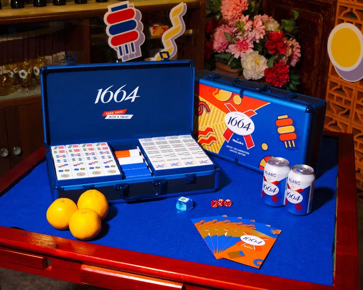 1664与法国艺术家Camille Walala联名限量版麻将套装展示图。画面中蓝色麻将礼盒打开陈列在桌面上，内含彩色设计麻将牌与筹码，盒身印有1664品牌标志与艺术家标志性几何图案，桌上还摆放1664 Blanc啤酒罐、柑橘与新年红包卡片，背景搭配节庆装饰与鲜花，整体风格活泼现代，呈现充满创意与趣味的新年礼物灵感。2026马年新年送礼推荐：拜年礼盒与新年礼物精选