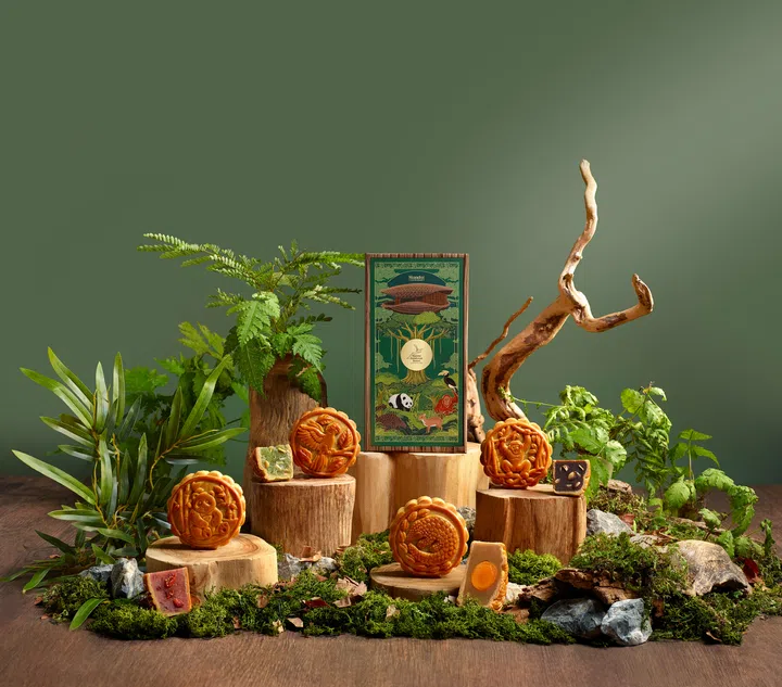Mandai Rainforest Resort by Banyan Tree 2026中秋月饼系列场景图，以森林绿背景和木桩、苔藓、石头及热带植物打造自然生态布景。画面中央陈列绿色四层月饼礼盒，盒面绘有Mandai Wildlife Reserve动物与森林元素；前景摆放四款传统烘焙月饼，表面分别压有熊猫、金刚鹦鹉、穿山甲及长臂猿等动物图案，并展示切开的月饼内馅，包括蛋黄与其他特色馅料。整体以野生动物与雨林为灵感，突出系列的自然主题与收藏感。