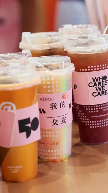《女友》50周年与At Tea联名活动现场，画面由三部分组成：左侧为多杯贴有《女友》50周年联名杯套的茶饮；中间两位参与者坐在粉色主题拍照装置前，手持趣味标语牌拍照留念；右侧展示“NÜYOU WHO CARES? WE CARE!”主题装置、粉色爱心气球及联名活动展示区，呈现联名活动温暖、轻松且充满互动感的氛围。