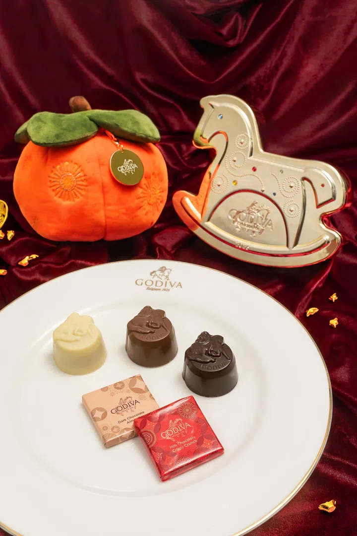 画面展示 GODIVA 马年新春巧克力系列陈列在酒红色丝绒布景上，整体氛围高雅且充满节庆感。背景左侧可见橙色吉祥橘子造型礼盒，顶部搭配绿色叶片装饰，并挂有 GODIVA 金色品牌吊牌；右侧则摆放银色木马造型巧克力礼盒，表面点缀彩色装饰元素，呼应马年主题。前景白色圆盘上整齐摆放多颗不同口味的 GODIVA 巧克力，包括白巧克力、牛奶巧克力与黑巧克力造型巧克力，旁边放有独立包装的小方块巧克力，包装以红金与粉金色调呈现新春图案。整体画面突显 GODIVA 以马年元素与吉祥寓意打造的新春巧克力礼盒，适合作为农历新年送礼与拜年分享的高质感甜点选择。2026马年年饼推荐：新春送礼与拜年点心精选
