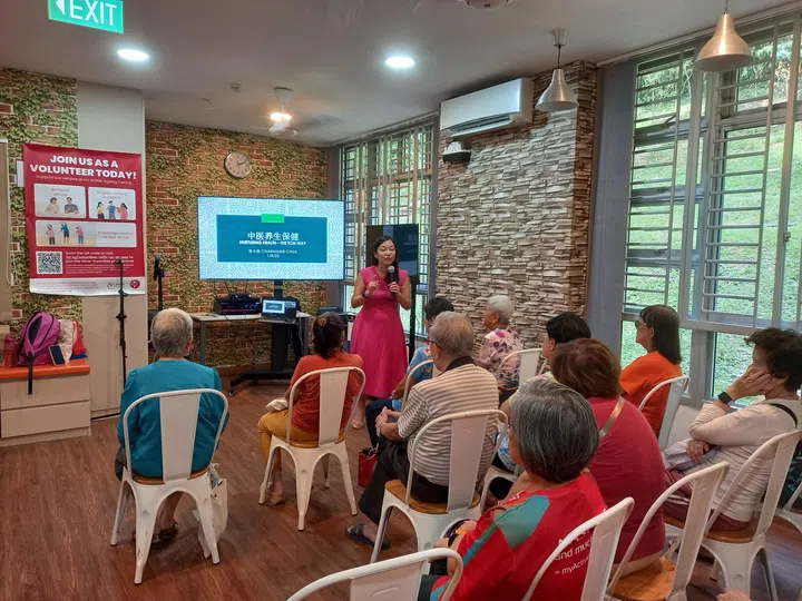 一位身穿粉色服装的讲师在教室前方进行中医养生讲座，台下的年长参与者专心聆听；背景萤幕上显示“中医养生·保健”的标题，现场气氛温馨而专注。哪里可以用SG Culture Pass？文化通行证活动推荐