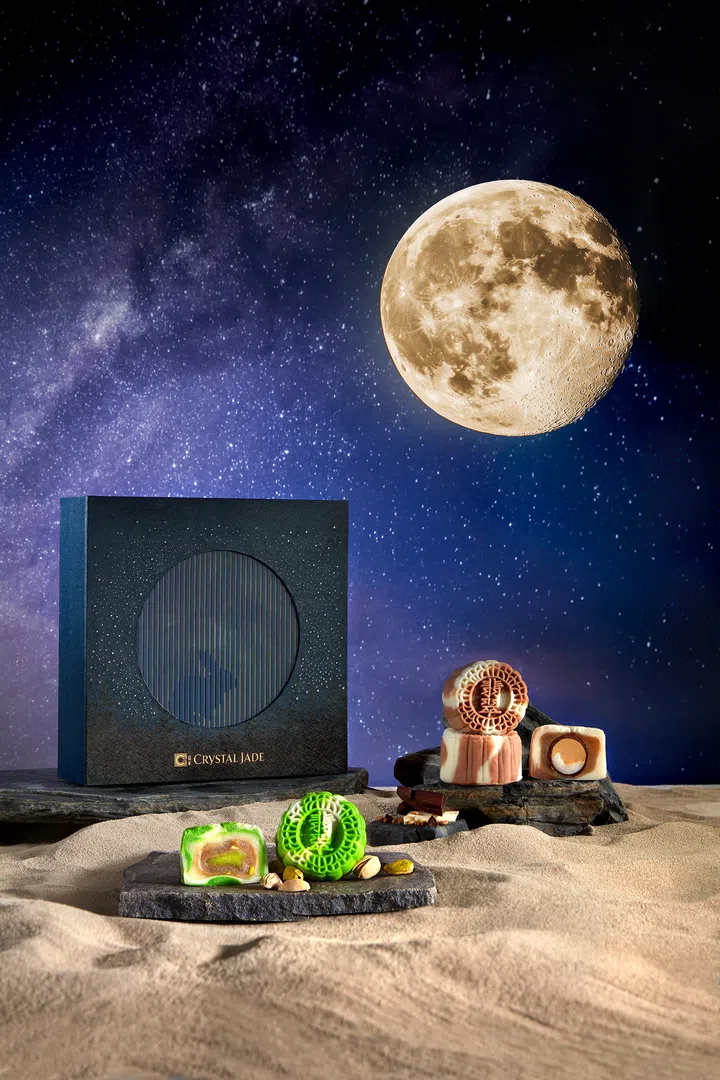 深蓝色的 Crystal Jade 星河主题礼盒置于沙面背景，背后点缀满天星辰与一轮皎洁圆月。前景摆放绿色与粉白相间的冰皮月饼，切开后可见内馅层次分明，周围点缀开心果与榛果，营造出梦幻科幻般的氛围。Crystal Jade，2025中秋月饼必买推荐。