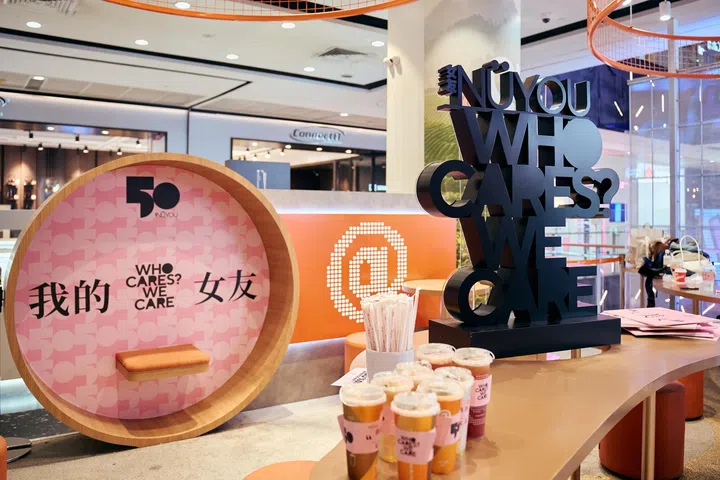 《女友》50周年与At Tea联名活动现场，左侧设有粉色圆形拍照打卡装置，印有“我的女友”及“WHO CARES? WE CARE.”字样；右侧摆放大型“NÜYOU WHO CARES? WE CARE.”立体主题装置，前方展示多杯贴有联名杯套的茶饮及活动物料，整体以粉色、橙色为主色调，营造温暖且充满互动感的活动空间。