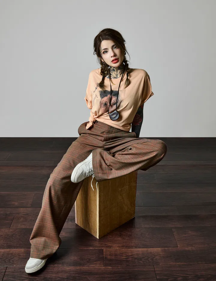 Casiline 坐在木箱上，穿着浅桃色 T 恤与宽松格纹长裤，脚踏白色球鞋。她扎着双辫子，露出颈部与手臂的纹身，身体微微前倾，表情自信而放松，背景是简单的灰墙与深色木地板。