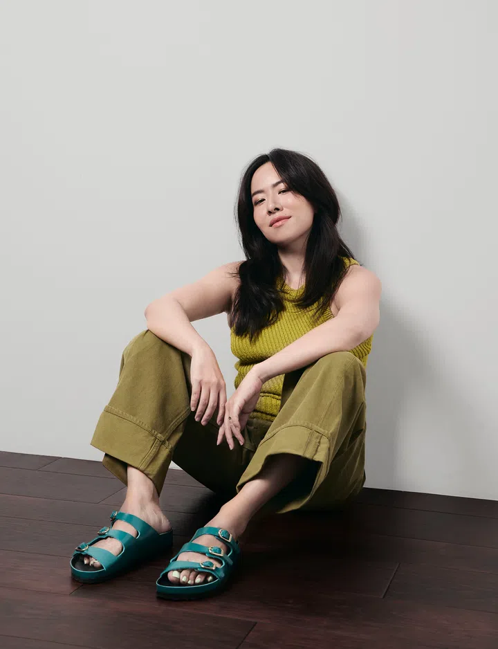 一名长发女子坐在深色木地板上，背靠浅灰色墙面，双腿自然弯曲，双手轻松搭放在膝盖上，面带淡淡微笑，自信且从容地望向镜头。她身穿黄绿色针织无袖上衣，搭配橄榄绿色宽版长裤，脚踩湖水蓝色三扣凉鞋，整体造型以大地色系与亮色鞋款形成鲜明对比，展现轻松自在又富有时尚感的风格。柔和自然光洒落于浅灰背景墙，为画面营造温暖、舒适且现代的氛围，整体呈现兼具亲和力与创意气息的人物肖像。