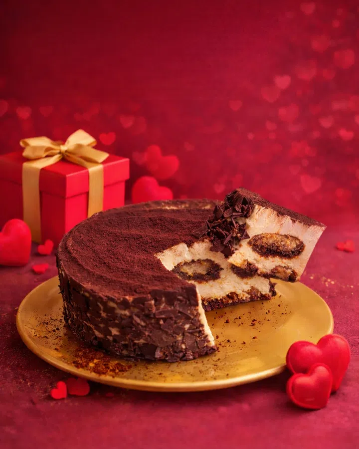 一款 Cat & the Fiddle Petite Tiramisu Cheesecake 放置在金色圆盘上，背景为红色渐层与爱心虚化图案，营造浓厚的情人节氛围。芝士蛋糕表面撒满细致的可可粉，侧边裹着深色巧克力碎片，呈现丰富层次感。蛋糕被切出一块并微微拉出，可清楚看见内部结构，由丝滑的芝士层与提拉米苏夹层组成，中间夹有咖啡浸润的蛋糕与浓郁巧克力内馅，质地细腻诱人。蛋糕上方点缀巧克力碎片，增加视觉与口感层次。背景左侧摆放一个红色礼盒，系着金色蝴蝶结，周围散落多个红色爱心装饰，与整体红色调相呼应，强化情人节送礼氛围。整张图片突显蛋糕作为情人节甜点礼物的精致与分享感。2026情人节礼物推荐：10款精致礼物让TA一秒心动