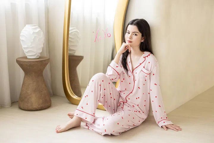 一名女性模特身穿粉色长袖长裤睡衣套装，坐在室内浅色木地板上，整体氛围简约而柔和。睡衣上布满细小红色爱心图案，并以红色滚边勾勒出领口、纽扣与袖口线条，设计俏皮又精致。模特轻靠在米色墙面旁，一手自然撑地，另一手托着下巴，神情从容。她身后是一面金色边框的落地镜，镜面上写着粉色“Xo Xo”字样，呼应情人节主题。镜子旁摆放着一张木质矮凳，上面放置白色雕塑感花瓶，为空间增添艺术气息。整体画面以粉色与米色为主调，呈现出舒适、轻松且带有情人节氛围的居家场景，突显爱心图案睡衣作为情人节礼物的精致与实用感。2026情人节礼物推荐：10款精致礼物让TA一秒心动