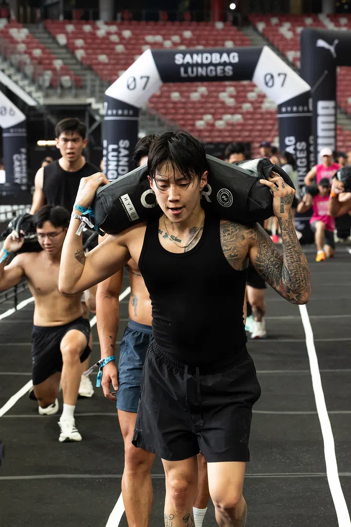 Jay Park在HYROX比赛中进行sandbag lunges负重弓步项目，将沙袋扛在肩上稳步前进，神情专注、身体发力明显，后方选手紧随其后，比赛气氛紧张而激烈。