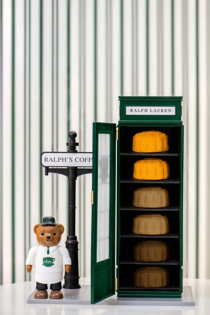 绿色 Ralph Lauren 标志的电话亭造型礼盒敞开，内部整齐摆放五枚迷你月饼。礼盒旁站立着身穿 Ralph’s Coffee 制服的 Polo Bear 公仔，背景为绿白条纹墙面，整体造型复古又俏皮。Ralph’s Coffee，2025中秋月饼必买推荐