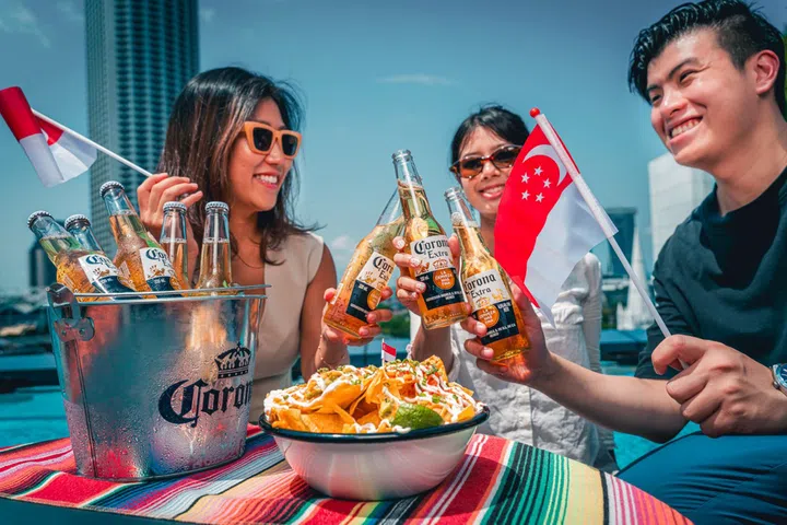 三名年轻人在Pullman Singapore Hill Street天台酒吧欢庆国庆，手持Corona啤酒举杯畅饮，并挥舞新加坡国旗，洋溢欢乐节庆氛围。桌上摆放一桶冰镇Corona啤酒及一盘墨西哥风味玉米片，背景可见蓝天与城市天际线，呈现适合与亲友相聚、享受美食美酒及欢庆国庆的高空餐饮体验。