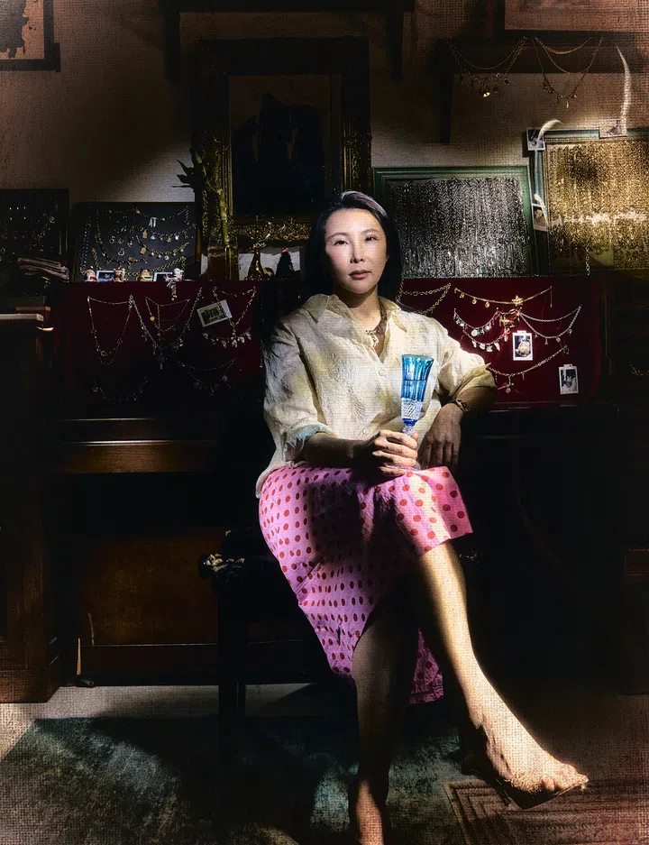 Echo Vintage Jewellery创办人Echo Wang坐在中古珠宝陈列前的全身肖像。她身穿米白色宽松暗纹衬衫、粉红色红圆点半裙与黑色尖头高跟鞋，双腿交叠，一手握着蓝色高脚玻璃杯，另一只手臂倚在身旁的深色木柜上，颈间及腕间搭配珠宝与腕表。身后的酒红色展示板与多个复古画框陈列着金银色链饰、吊坠和小幅照片，周围可见雕花镜框、绿植及装饰摆件。斜落的光线照亮她的脸庞、衣裙与小腿，衬托出木质家具、深色地毯和珠宝陈列的细节，整张照片呈现带有颗粒与织纹质感的怀旧色调。
