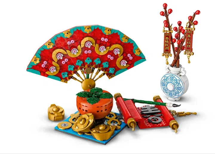 画面展示 LEGO Good Fortune「五福迎春」（80117）积木套装的多个组合摆件。左侧是一把展开的中式折扇造型积木，扇面以红色为主，搭配金色、绿色与粉色花纹装饰，呈现对称几何与花卉图案。扇子底座前方摆放多件金色元宝造型积木，以及一颗橘色柿子摆件，象征吉祥与丰收。右侧可见一组白蓝色花瓶造型积木，插着红色果实枝条，并悬挂红底金字的新春吊饰与对联挂件。前方还摆放一组红色卷轴造型积木，呈现传统书法卷轴结构。整体配色以红、金、绿与蓝白为主，融合传统农历新年元素与积木结构设计，适合作为居家空间的新春装饰摆设与收藏展示。