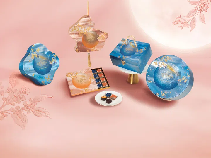 GODIVA 2026中秋节限定巧克力系列礼盒场景图，以柔和粉色背景与圆月意象营造节庆氛围。画面展示多款蓝色及粉金色GODIVA中秋礼盒，包括圆形、方形与不规则造型包装，表面饰有金色花卉与月夜图案。中央打开的礼盒可见多款独立包装巧克力，前方白色小碟上摆放巧克力、迷你挞及巧克力方片，整体呈现精致华丽的中秋送礼氛围。