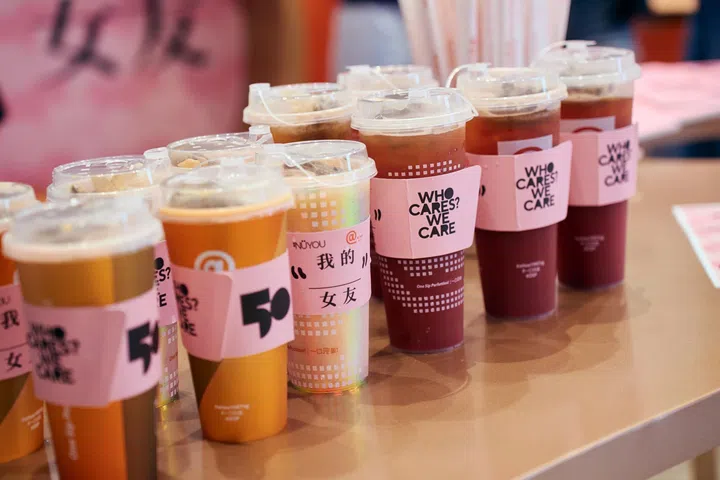 多杯《女友》与At Tea联名推出的限定饮品“女友莓果伯爵”整齐摆放于桌面，包括“晴天版”与“梦幻版”两种版本。饮品套上粉色联名杯套，印有《女友》50周年标志、“WHO CARES? WE CARE.”及“我的女友”等主题设计，呼应联名合作与“CARE”主题。
