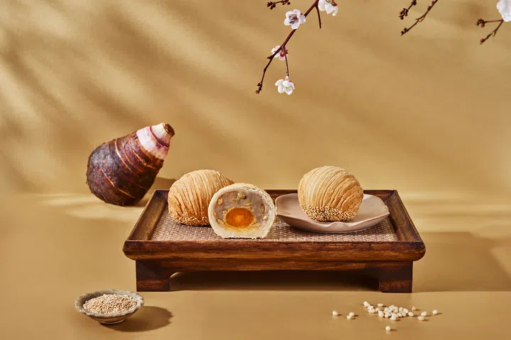 木质托盘上摆放三枚 Paradise Group 潮州单黄芋泥酥，其中一枚对半切开，露出细腻的芋泥馅与金黄咸蛋黄。背景摆有完整芋头与芝麻点缀，衬托出食材本味。画面整体以暖金色为基调，配以梅花枝点缀，呈现典雅氛围。Paradise Group，2025中秋月饼必买推荐