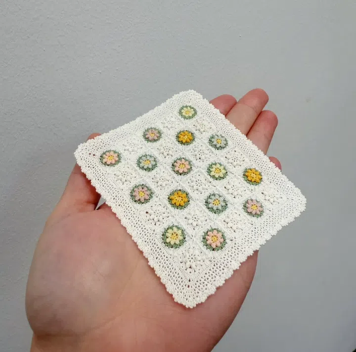一只手掌心托着一块微钩编作品，呈矩形白色桌巾，上面排列着黄、粉、绿花纹的圆形图案，展现精细的针线工艺。smolchet Marilyn亲手制作的迷你装饰桌巾。