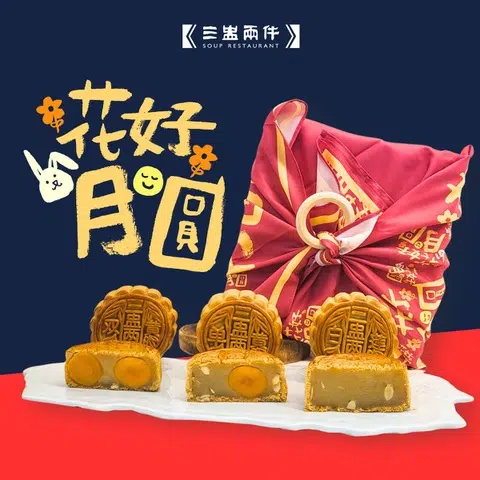 三盅两件月饼礼盒以红色印花布包裹，系上圆环结扣，充满传统风韵。前方白色瓷盘上摆放三枚金黄烘焙月饼，其中一枚切开，露出双黄白莲蓉馅料。背景搭配手绘风格的“花好月圆”文字与小兔插画，节日氛围浓厚。Soup Restaurant，2025中秋月饼必买推荐。