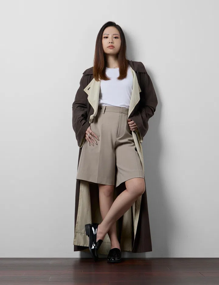 室内时尚人像照，一名女性站在简约的浅灰白色墙面前拍摄全身照，地面为深色木纹地板。她留着中长直发，妆容自然精致，神情沉稳自信，直视镜头。身穿白色圆领上衣，外搭深棕色长版风衣，内里为浅米色衬里；下身搭配卡其色高腰西装短裤，并穿着黑色亮面乐福鞋。她一脚微微交叉站立，双手自然插在风衣口袋边，整体造型利落现代，呈现干练而优雅的都会女性形象。