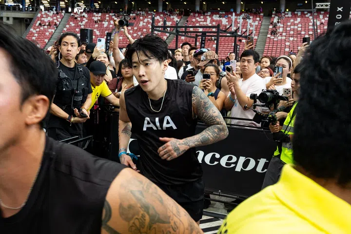 Jay Park在HYROX比赛中奔跑进入下一个训练项目，神情专注略显疲惫，身穿AIA运动背心，周围观众举起手机围观拍摄，现场气氛紧张而热烈。