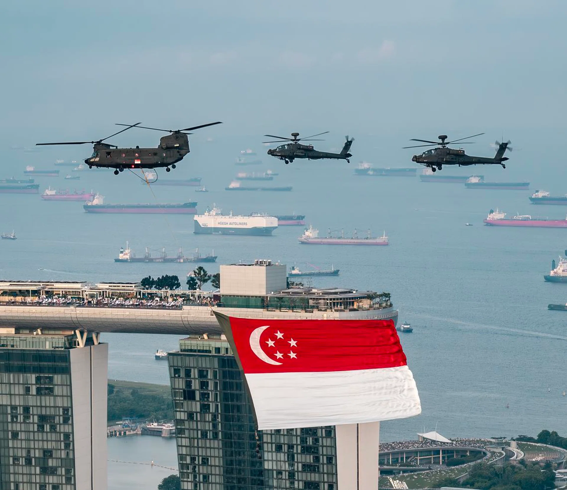 新加坡滨海湾（Marina Bay）上空，三架新加坡空军军用直升机编队飞行，其中一架CH-47支奴干直升机悬挂着一面巨大的新加坡国旗，旗帜迎风展开，鲜红与白色在空中格外醒目。前景可见滨海湾金沙酒店（Marina Bay Sands）标志性的三座塔楼，国旗正从其中一座塔楼前方掠过。远处的新加坡港口布满数十艘货轮与集装箱船，平静的海面延伸至地平线，天空带有淡淡薄雾，营造出庄严而壮观的氛围。整幅画面展现新加坡国庆庆典彩排期间的空中飞行表演，同时凸显这座海港城市的天际线与繁忙港口景观。