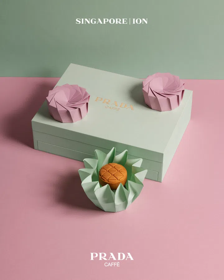 浅绿色礼盒上印有金色 “PRADA Caffè” 字样，礼盒旁与顶部分别摆放折纸花瓣造型的月饼包装。下方一枚月饼置于绿色折纸托中，宛如花朵盛开，整体以粉色与绿色背景衬托，呈现简洁时尚的视觉效果。Prada Caffè，2025中秋月饼必买推荐