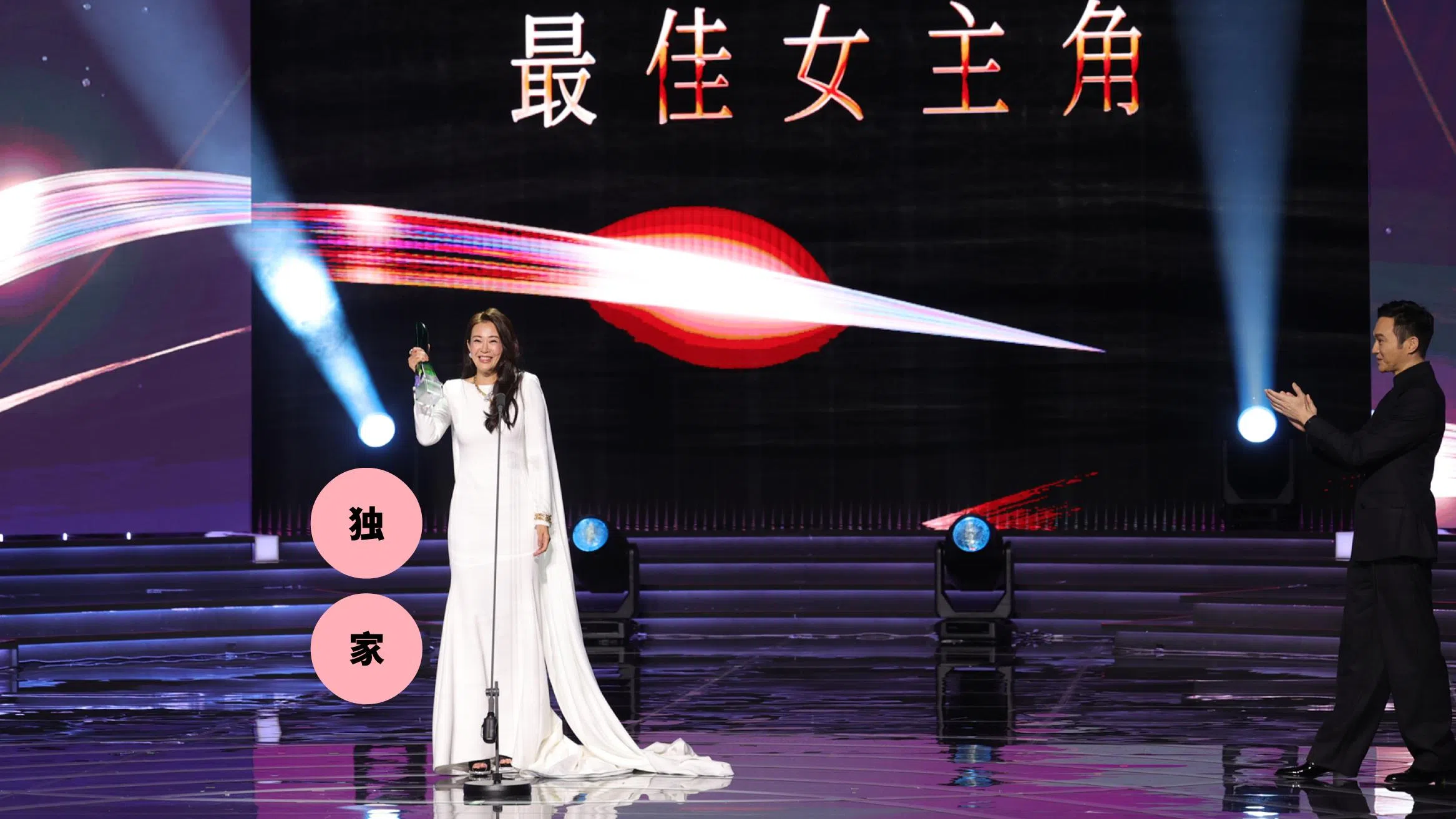 这是一张在《红星大奖》颁奖典礼现场拍摄的照片。舞台中央，刘子绚身穿一袭简洁优雅的白色长礼服，裙摆自然拖地，站在麦克风前，手中举着最佳女主角奖座，脸上带着开心又略显感动的笑容。她的整体造型端庄大方，气质沉稳，成为全场焦点。
舞台背景以黑色为主，上方大字写着“最佳女主角”，搭配红色与白色光影线条，营造出隆重而充满张力的视觉效果。舞台灯光从两侧打下，形成聚光效果，强调得奖者的重要时刻。
画面右侧，一位男艺人身穿黑色西装，正面向刘子绚鼓掌，神情带着欣赏与祝贺。舞台地面呈现镜面反射效果，映出灯光与人物轮廓，让整体画面更具层次感与仪式感。
画面左侧叠加了两个粉色圆形图形，分别写着“独”“家”字样，显示为媒体内容的视觉设计元素。整体画面捕捉了刘子绚获奖当下的荣耀瞬间，气氛温暖而充满掌声与肯定。刘子绚首夺视后：22年7次提名终圆梦，她把时间走成答案
