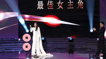 这是一张在《红星大奖》颁奖典礼现场拍摄的照片。舞台中央，刘子绚身穿一袭简洁优雅的白色长礼服，裙摆自然拖地，站在麦克风前，手中举着最佳女主角奖座，脸上带着开心又略显感动的笑容。她的整体造型端庄大方，气质沉稳，成为全场焦点。
舞台背景以黑色为主，上方大字写着“最佳女主角”，搭配红色与白色光影线条，营造出隆重而充满张力的视觉效果。舞台灯光从两侧打下，形成聚光效果，强调得奖者的重要时刻。
画面右侧，一位男艺人身穿黑色西装，正面向刘子绚鼓掌，神情带着欣赏与祝贺。舞台地面呈现镜面反射效果，映出灯光与人物轮廓，让整体画面更具层次感与仪式感。
画面左侧叠加了两个粉色圆形图形，分别写着“独”“家”字样，显示为媒体内容的视觉设计元素。整体画面捕捉了刘子绚获奖当下的荣耀瞬间，气氛温暖而充满掌声与肯定。刘子绚首夺视后：22年7次提名终圆梦，她把时间走成答案