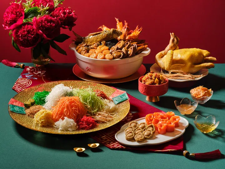农历新年外带年菜与捞起鱼生摆盘展示图，前景为金色大盘盛装的捞起鱼生，铺满白萝卜丝、红萝卜丝、青萝卜丝、酥脆配料与酱包，周围搭配鲍鱼片与三文鱼片；背景中央是一锅丰盛盆菜，内含整条鱼、鲜虾、鲍鱼、带子与香菇等海味食材，右侧摆放整只白切鸡与人参，桌面点缀红色年节布饰、金元宝装饰与花卉，整体营造热闹喜庆的新加坡农历新年团圆饭与外卖年菜氛围。