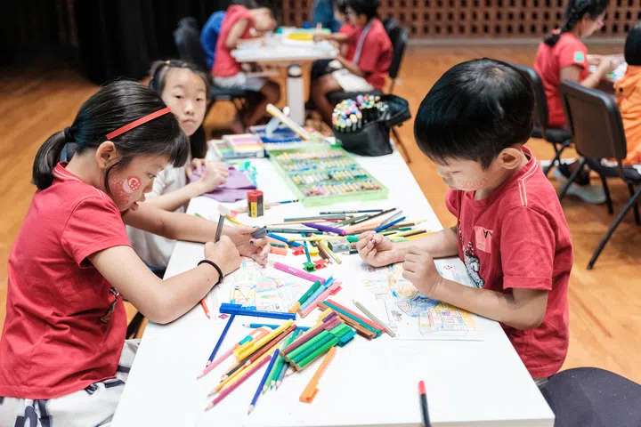 多名小朋友参加National Museum of Singapore举办的填色活动，围坐在桌前专注为图画上色。桌面摆满彩色铅笔、彩色笔及绘画工具，孩子们认真创作属于自己的作品，部分小朋友脸上还绘有国庆主题彩绘。现场洋溢轻松欢乐的氛围，展现适合亲子参与的创意艺术体验。