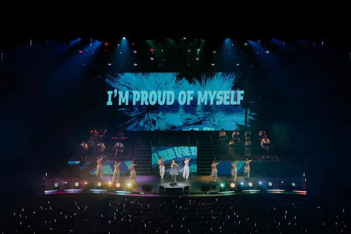 DIOR大颖在新加坡演唱会大型舞台上表演，站在舞台中央，与多位舞者同台演出，后方乐队同步演奏。舞台灯光呈现多色效果，LED大屏幕显示“I’M PROUD OF MYSELF”字样，台下观众挥舞应援灯，营造出热烈且充满能量的现场氛围。